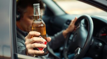 A drunk driver behind the wheel of his car holding a beer in his hand.